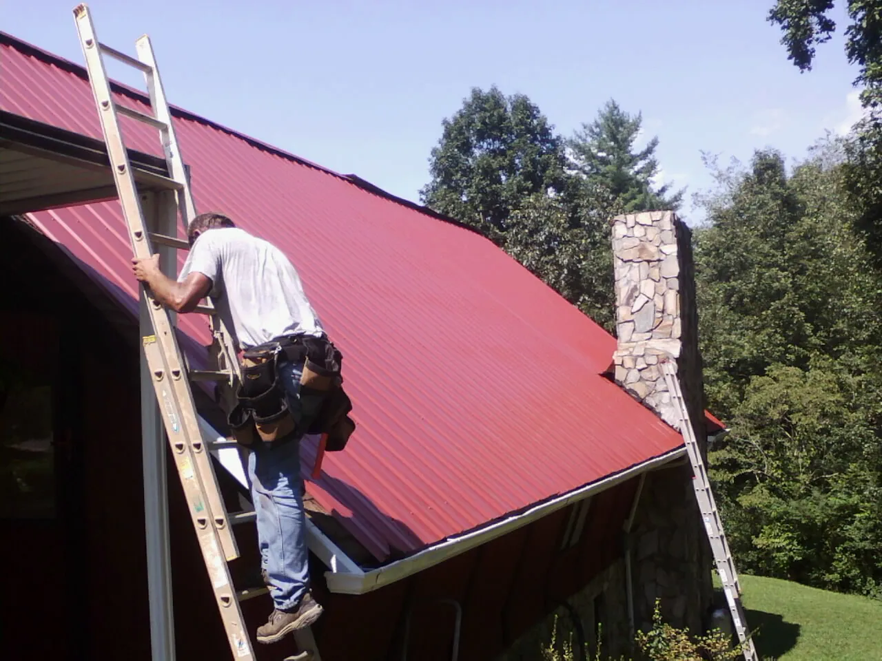 Roofer on a ladder beside a red metal roof and stone chimney — Soffit & Fascia Repair by Roofing Brothers in Austin